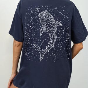 May include: Navy blue t-shirt featuring a white illustration of a whale shark on the back, surrounded by a starry design. The shirt has short sleeves and a relaxed fit. The word "NAVY" is printed on the bottom right.