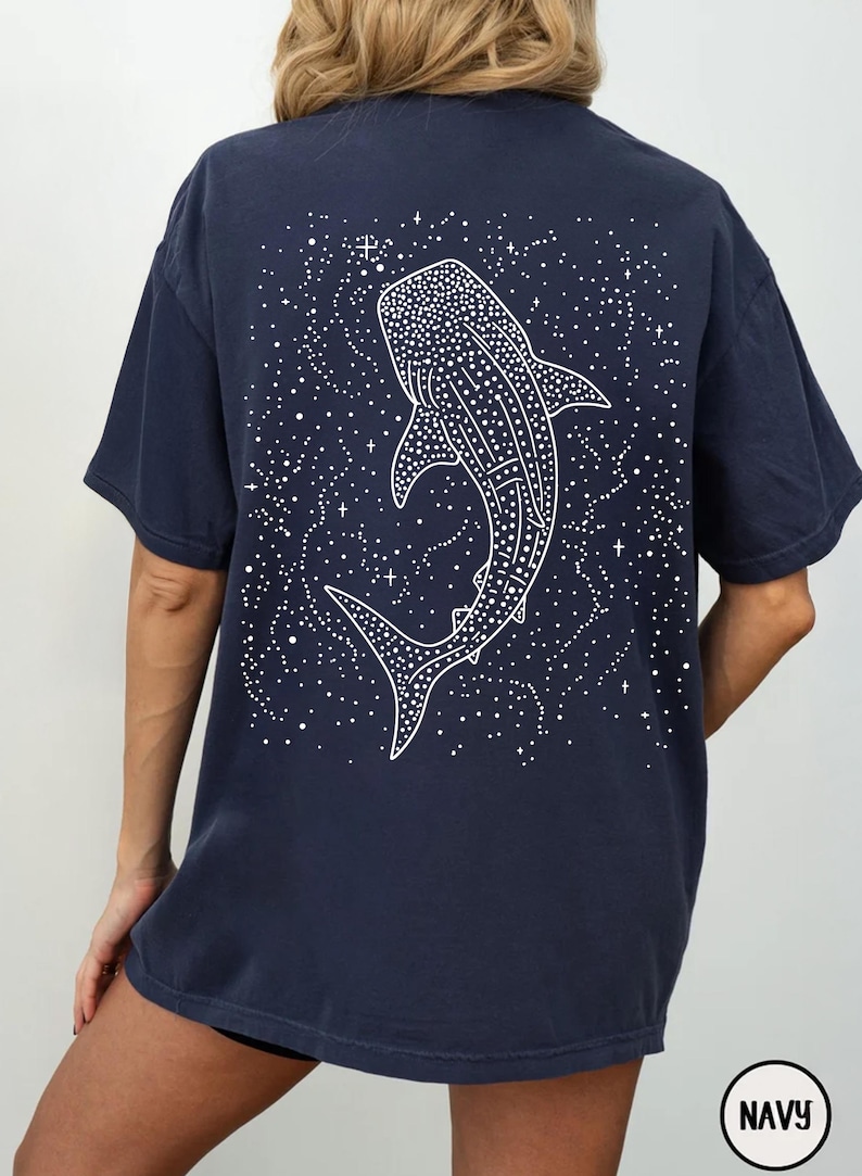 May include: Navy blue t-shirt featuring a white illustration of a whale shark on the back, surrounded by a starry design. The shirt has short sleeves and a relaxed fit. The word "NAVY" is printed on the bottom right.