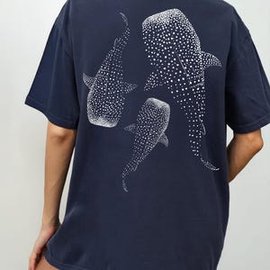 May include: Navy blue t-shirt featuring a white illustration of three whale sharks. The whale sharks are depicted with white spots. The shirt has short sleeves and a round neckline. The word "NAVY" is in a white circle in the bottom right corner.