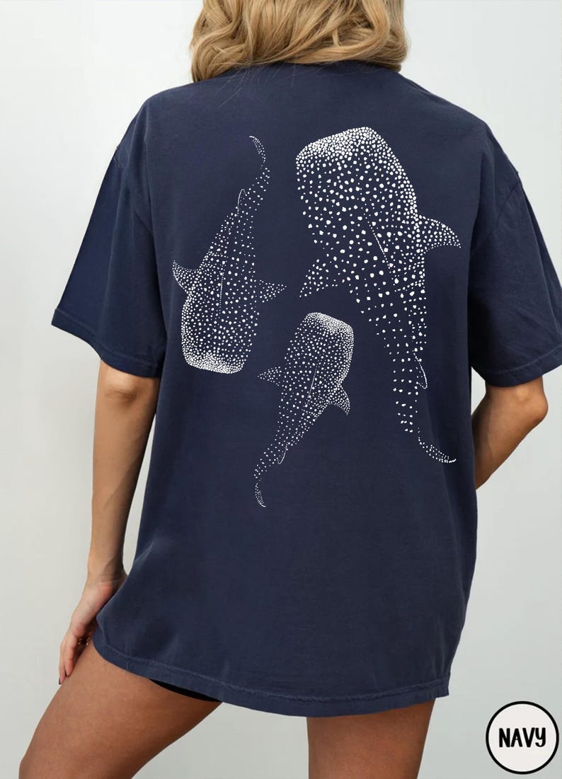 May include: Navy blue t-shirt featuring a white illustration of three whale sharks. The whale sharks are depicted with white spots. The shirt has short sleeves and a round neckline. The word "NAVY" is in a white circle in the bottom right corner.
