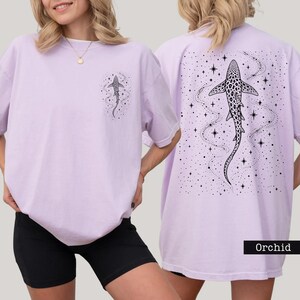 Puede incluir: Camiseta color orqu&iacute;dea con un gr&aacute;fico de tibur&oacute;n en la parte delantera y trasera. La parte delantera presenta un peque&ntilde;o dise&ntilde;o de tibur&oacute;n, mientras que la parte trasera muestra una ilustraci&oacute;n de tibur&oacute;n m&aacute;s grande con un fondo estrellado. La camiseta es de corte holgado.
