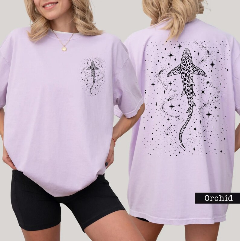 Puede incluir: Camiseta color orqu&iacute;dea con un gr&aacute;fico de tibur&oacute;n en la parte delantera y trasera. La parte delantera presenta un peque&ntilde;o dise&ntilde;o de tibur&oacute;n, mientras que la parte trasera muestra una ilustraci&oacute;n de tibur&oacute;n m&aacute;s grande con un fondo estrellado. La camiseta es de corte holgado.