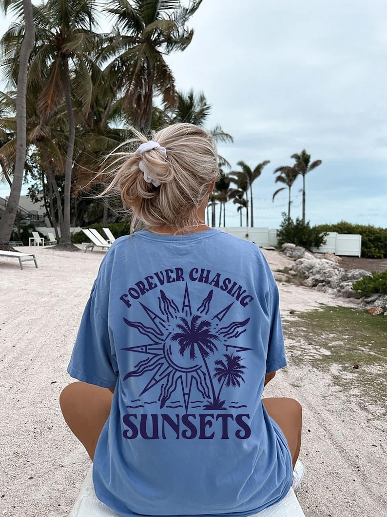 Puede incluir: Una camiseta azul claro con un gr&aacute;fico de un sol, palmeras y el texto "FOREVER CHASING SUNSETS" impreso en la espalda.