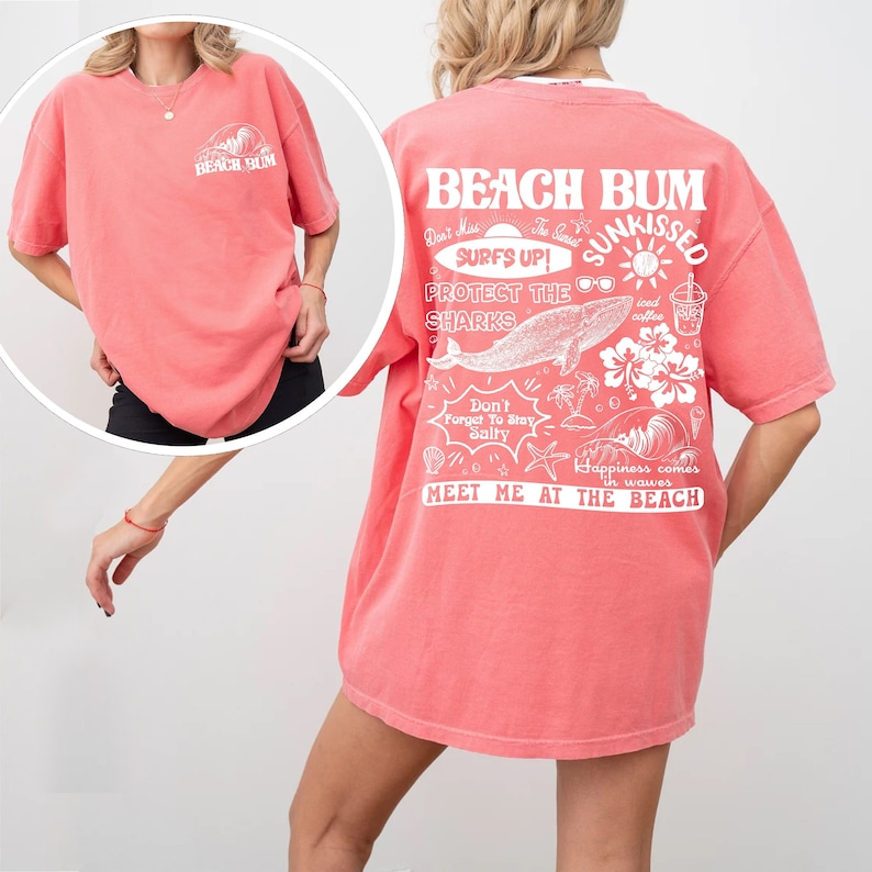 May include: Coral-coloured t-shirt with white graphic text and illustrations. The front features "Beach Bum" text and a wave design. The back displays "Beach Bum," "Surfs Up!," a whale, and other beach-themed graphics. The shirt has short sleeves.