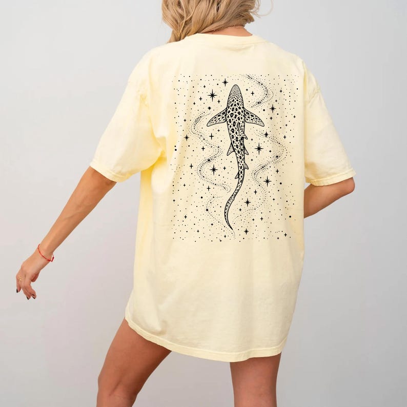 Puede incluir: Camiseta amarillo p&aacute;lido con una ilustraci&oacute;n en blanco y negro de un tibur&oacute;n con un patr&oacute;n de puntos y una cola larga. El dise&ntilde;o est&aacute; rodeado de estrellas y l&iacute;neas onduladas. La camiseta tiene mangas cortas y un corte holgado.