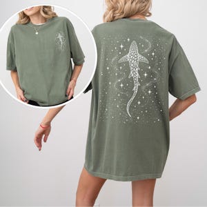 Puede incluir: Camiseta verde oliva con un dise&ntilde;o de tibur&oacute;n ballena blanco en la espalda y un dise&ntilde;o a juego m&aacute;s peque&ntilde;o en la parte delantera. El tibur&oacute;n ballena est&aacute; rodeado de estrellas y un patr&oacute;n arremolinado. La camiseta tiene mangas cortas y un corte holgado.