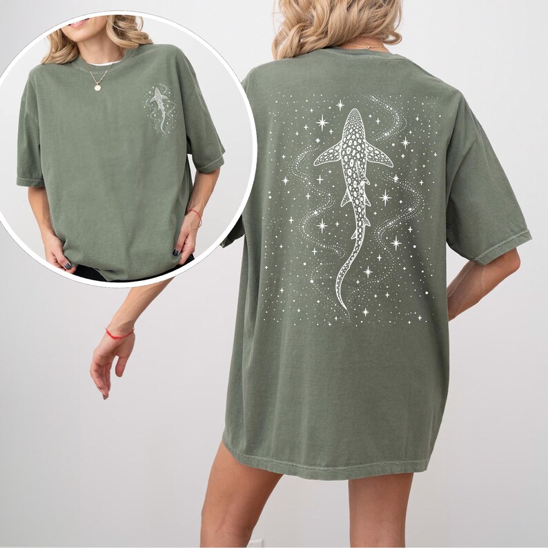 Puede incluir: Camiseta verde oliva con un dise&ntilde;o de tibur&oacute;n ballena blanco en la espalda y un dise&ntilde;o a juego m&aacute;s peque&ntilde;o en la parte delantera. El tibur&oacute;n ballena est&aacute; rodeado de estrellas y un patr&oacute;n arremolinado. La camiseta tiene mangas cortas y un corte holgado.