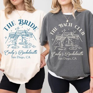 May include: Two t-shirts with a bachelorette party theme. The white t-shirt says "The Bride" with a design of four martini glasses and the text "Emily's Bachelorette, San Diego, CA". The gray t-shirt says "The Bach Club" with a design of four martini glasses and the text "Emily's Bachelorette, San Diego, CA".