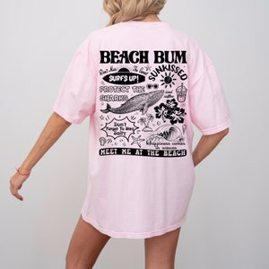 May include: A pale pink t-shirt featuring a black and white beach-themed graphic on the back. The design includes the words "Beach Bum," a whale, and other illustrations. The phrase "Meet Me at the Beach" is also visible.