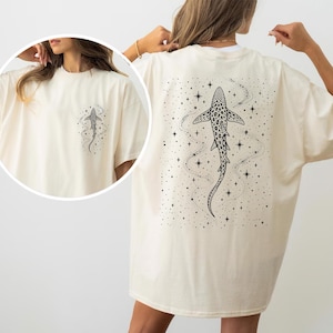 Puede incluir: Camiseta color crema con un dise&ntilde;o de tibur&oacute;n en blanco y negro en la espalda y un gr&aacute;fico de tibur&oacute;n m&aacute;s peque&ntilde;o en la parte delantera. El dise&ntilde;o del tibur&oacute;n incluye estrellas y l&iacute;neas punteadas, creando un tema celestial. La camiseta tiene un corte holgado.