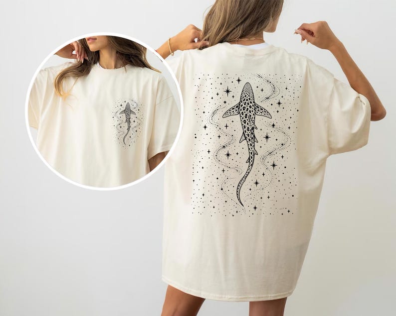 Puede incluir: Camiseta color crema con un dise&ntilde;o de tibur&oacute;n en blanco y negro en la espalda y un gr&aacute;fico de tibur&oacute;n m&aacute;s peque&ntilde;o en la parte delantera. El dise&ntilde;o del tibur&oacute;n incluye estrellas y l&iacute;neas punteadas, creando un tema celestial. La camiseta tiene un corte holgado.