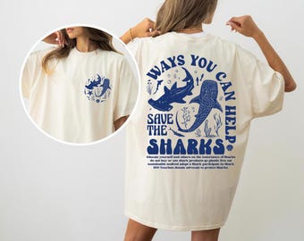 May include: A cream coloured t-shirt with a blue graphic on the back that says "Ways You Can Help Save The Sharks". The graphic features two sharks and seaweed. The front of the shirt has a smaller blue graphic of a shark and seaweed.