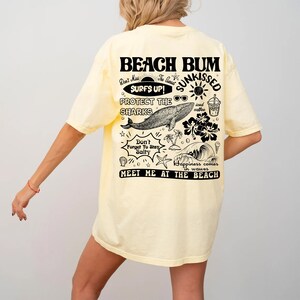 May include: A pale yellow t-shirt featuring a beach-themed graphic on the back. The design includes the words "BEACH BUM" and other illustrations and phrases such as "SURFS UP!" and "MEET ME AT THE BEACH."