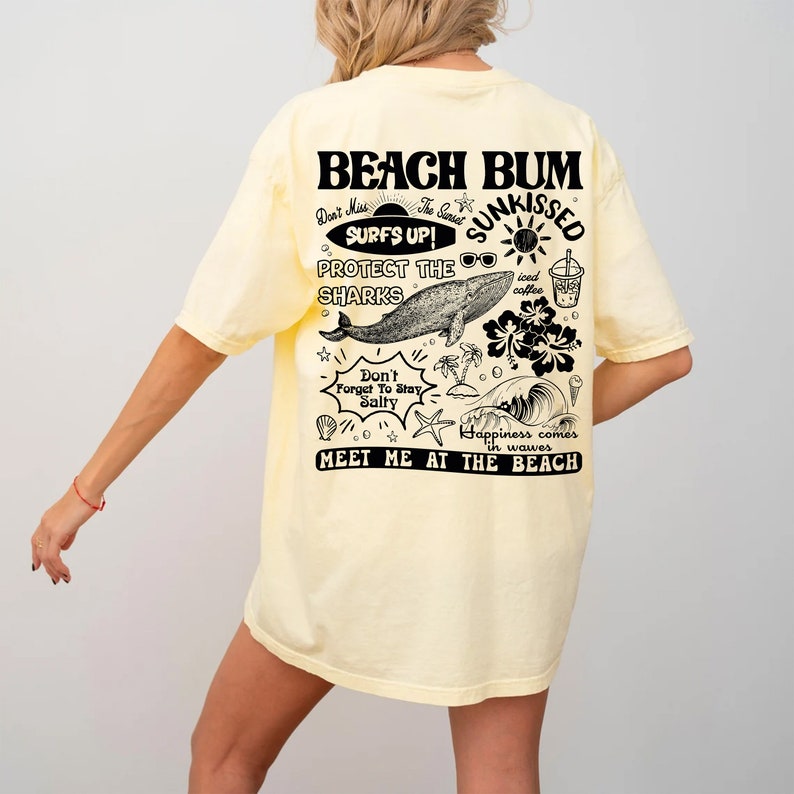 May include: A pale yellow t-shirt featuring a beach-themed graphic on the back. The design includes the words "BEACH BUM" and other illustrations and phrases such as "SURFS UP!" and "MEET ME AT THE BEACH."