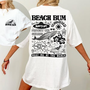 May include: White oversized t-shirt featuring a black graphic on the back with the words "Beach Bum" and beach-themed illustrations. The front of the shirt has a small graphic of a wave and the words "Beach Bum".