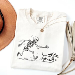 May include: An off-white short-sleeve t-shirt featuring a black ink graphic of a running skeleton missing an arm, chasing a small dog that is carrying a bone in its mouth. The shirt has a Comfort Colors brand label at the inner neckline.