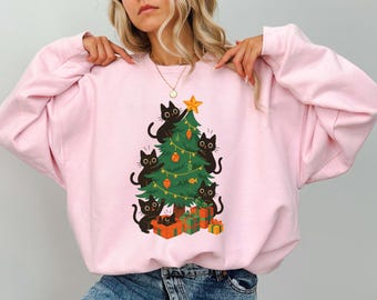 Op de afbeelding: Een lichtroze sweatshirt met een kerstboomontwerp. De boom is versierd met zwarte katten, ballen en een ster. Zwarte katten beklimmen de boom, met cadeaus aan de basis. Een leuk, feestelijk ontwerp.
