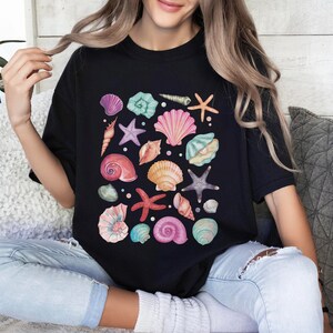 K&ouml;nnte beinhalten: Ein schwarzes T-Shirt mit einem farbenfrohen Design aus Muscheln und Seesternen. Das Design zeigt eine Vielzahl von Muscheln in den Farben Rosa, Blau, Gr&uuml;n und Orange sowie Seesterne in den Farben Rosa und Lila.