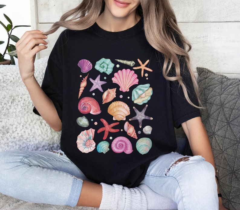 K&ouml;nnte beinhalten: Ein schwarzes T-Shirt mit einem farbenfrohen Design aus Muscheln und Seesternen. Das Design zeigt eine Vielzahl von Muscheln in den Farben Rosa, Blau, Gr&uuml;n und Orange sowie Seesterne in den Farben Rosa und Lila.