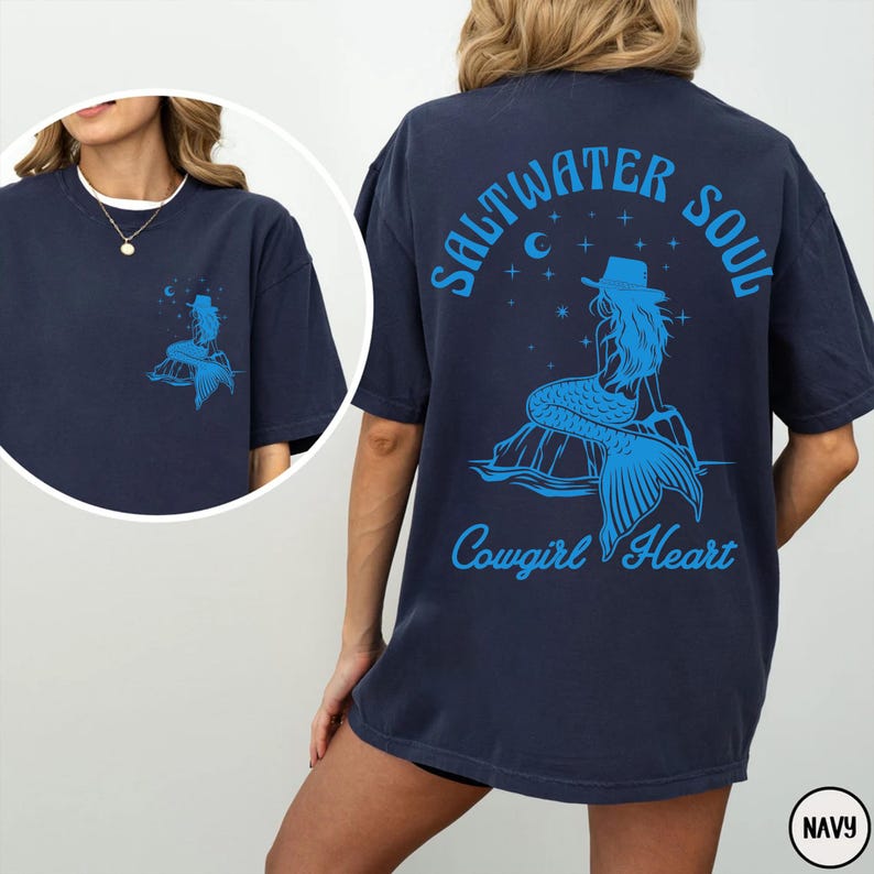 Peut inclure: Un t-shirt bleu marine avec un motif de sir&egrave;ne portant un chapeau de cow-boy et le texte "Saltwater Soul Cowgirl Heart".