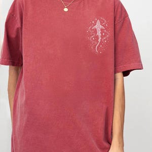 Puede incluir: Camiseta de color carmes&iacute; con un gr&aacute;fico blanco de un pez y estrellas en la parte superior izquierda del pecho. La camiseta tiene mangas cortas y cuello redondo. La palabra "Crimson" est&aacute; en la esquina inferior derecha.