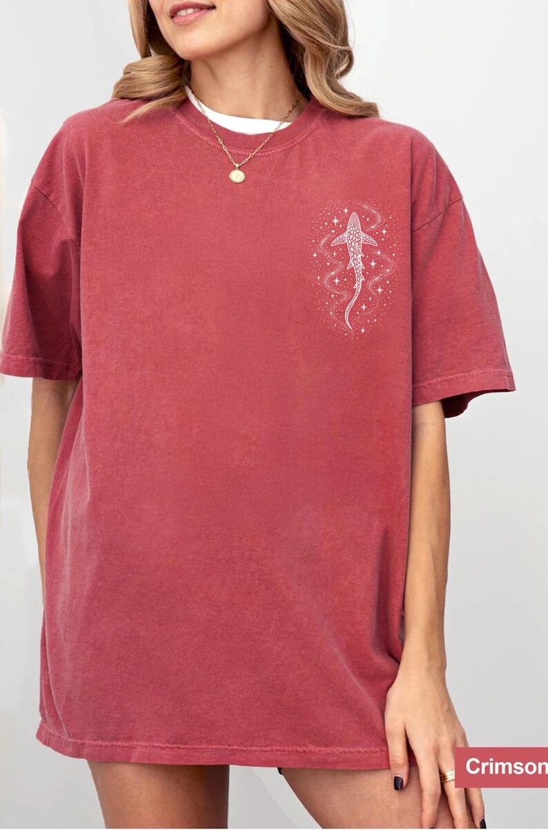 Puede incluir: Camiseta de color carmes&iacute; con un gr&aacute;fico blanco de un pez y estrellas en la parte superior izquierda del pecho. La camiseta tiene mangas cortas y cuello redondo. La palabra "Crimson" est&aacute; en la esquina inferior derecha.