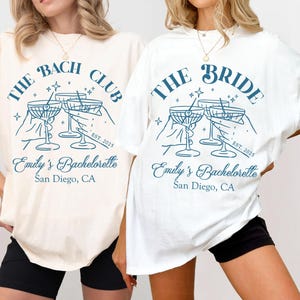 May include: Two white t-shirts with a blue graphic of a group of hands holding up martini glasses. The text on the shirts reads "The Bach Club" and "The Bride" with the text "Emily's Bachelorette San Diego, CA" below the graphic. The shirts are on a white background.