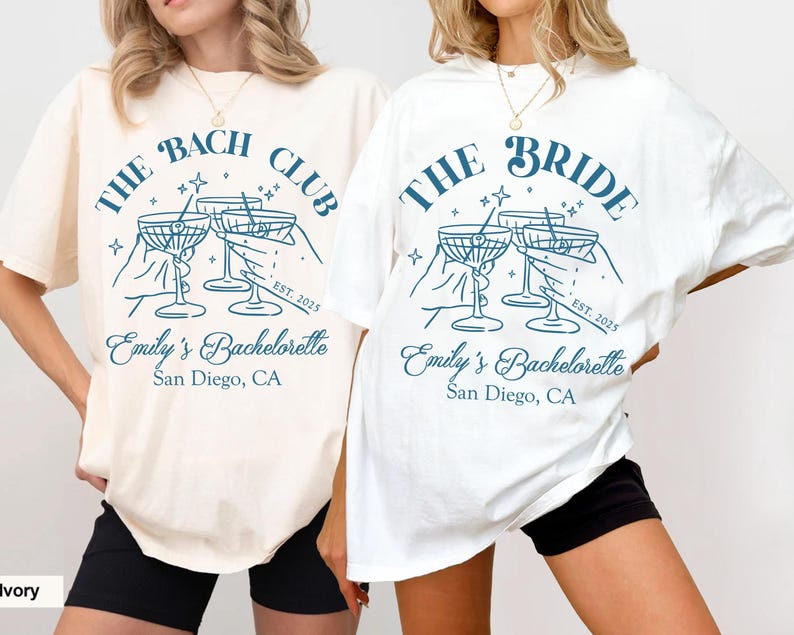 May include: Two white t-shirts with a blue graphic of a group of hands holding up martini glasses. The text on the shirts reads "The Bach Club" and "The Bride" with the text "Emily's Bachelorette San Diego, CA" below the graphic. The shirts are on a white background.
