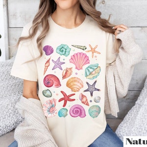 K&ouml;nnte beinhalten: Ein wei&szlig;es T-Shirt mit einem farbenfrohen Design aus Muscheln und Seesternen. Das Design zeigt eine Vielzahl von Muscheln in den Farben Rosa, Lila, Blau und Gr&uuml;n sowie Seesterne und andere maritime Elemente.