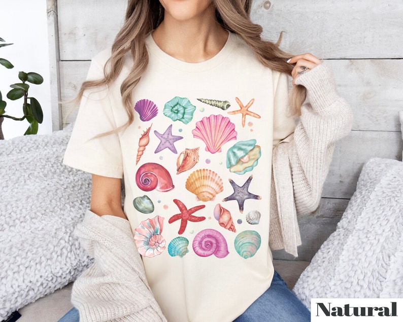K&ouml;nnte beinhalten: Ein wei&szlig;es T-Shirt mit einem farbenfrohen Design aus Muscheln und Seesternen. Das Design zeigt eine Vielzahl von Muscheln in den Farben Rosa, Lila, Blau und Gr&uuml;n sowie Seesterne und andere maritime Elemente.