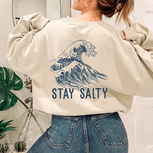 May include: A cream-colored sweatshirt with a blue wave graphic and the text "STAY SALTY" printed on the back. The sweatshirt is worn by a person with long brown hair and is paired with blue jeans.