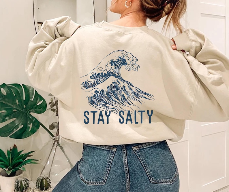 May include: A cream-colored sweatshirt with a blue wave graphic and the text "STAY SALTY" printed on the back. The sweatshirt is worn by a person with long brown hair and is paired with blue jeans.