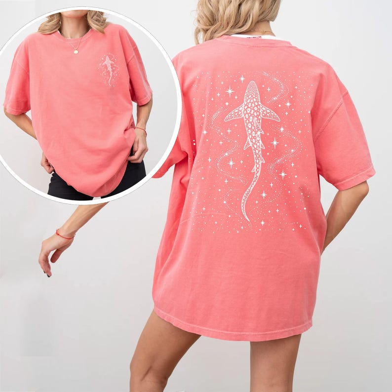 Puede incluir: Camiseta de color coral con un dise&ntilde;o de tibur&oacute;n blanco en la espalda y un dise&ntilde;o m&aacute;s peque&ntilde;o en la parte delantera. El gr&aacute;fico del tibur&oacute;n est&aacute; rodeado de estrellas y l&iacute;neas onduladas. La camiseta tiene mangas cortas y un corte holgado.