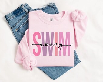 Peut inclure: Un sweat-shirt rose clair avec le mot "SWIM" en grandes lettres roses et violettes, avec le nom "avery" en cursive en dessous. Le sweat-shirt est présenté sur un jean bleu.