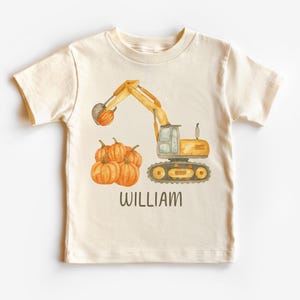 May include: Cream-colored toddler t-shirt featuring a watercolor illustration of a yellow excavator lifting a pumpkin, with a pile of pumpkins below. The name "WILLIAM" is printed in brown letters below the pumpkins.