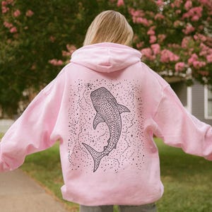 May include: A light pink hoodie featuring a black and white whale shark design on the back. The whale shark is surrounded by a constellation of dots and stars. The hoodie has a hood and long sleeves.