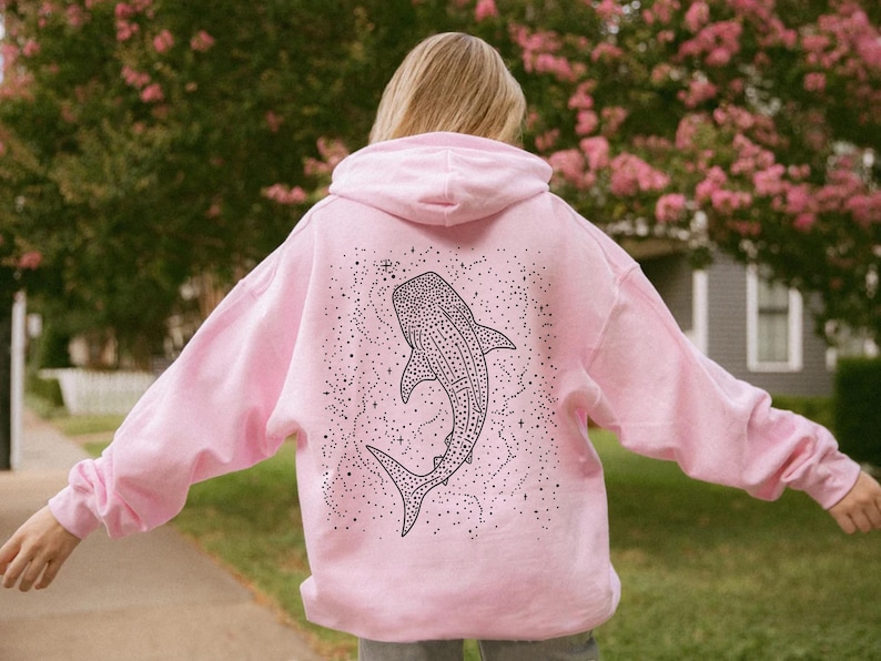 May include: A light pink hoodie featuring a black and white whale shark design on the back. The whale shark is surrounded by a constellation of dots and stars. The hoodie has a hood and long sleeves.