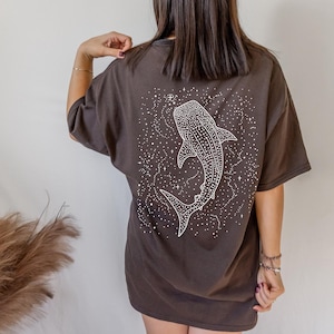 May include: A dark brown t-shirt featuring a white illustration of a whale shark surrounded by stars. The graphic is on the back of the shirt. The shirt has short sleeves and a relaxed fit.