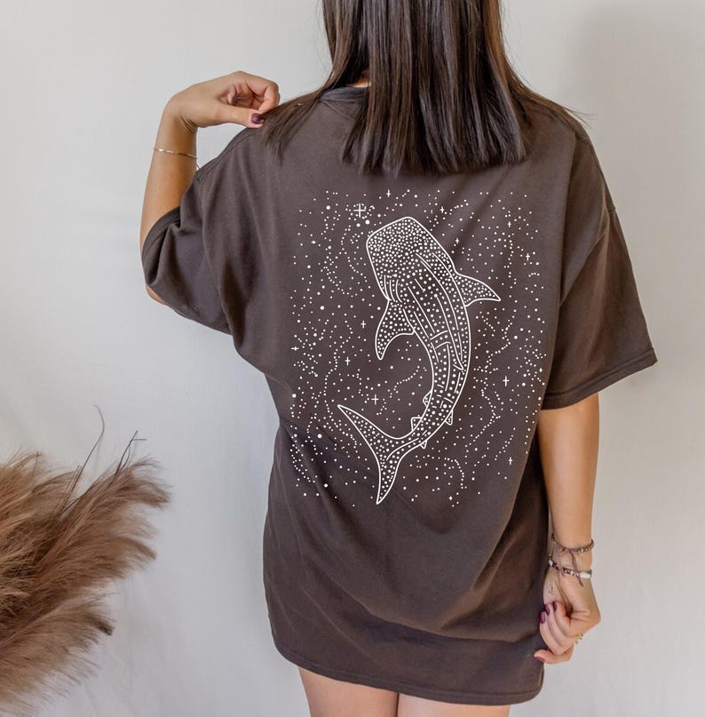 May include: A dark brown t-shirt featuring a white illustration of a whale shark surrounded by stars. The graphic is on the back of the shirt. The shirt has short sleeves and a relaxed fit.