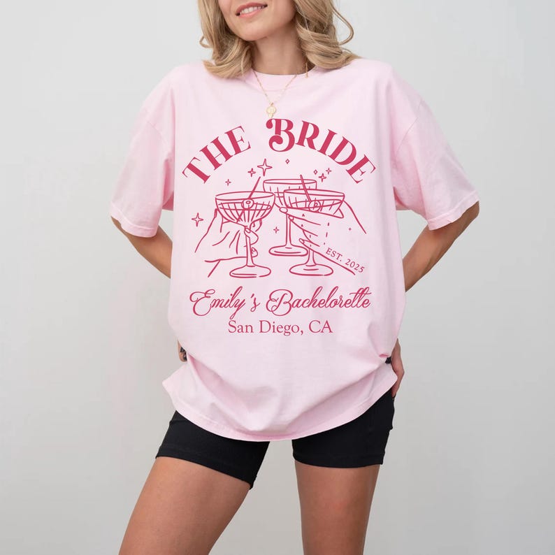 May include: Pink t-shirt with the text "The Bride" in red lettering, featuring a graphic of three hands holding up champagne glasses. The text "Emily's Bachelorette" and "San Diego, CA" are also printed in red.