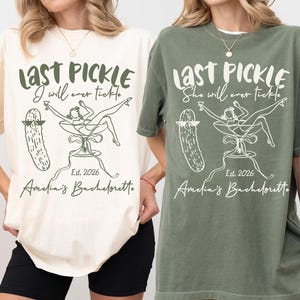 May include: Two t-shirts, one cream and one olive green, with a graphic design. The design includes the text "LAST PICKLE" and "Amelia's Bachelorette" with a pickle and a woman in a martini glass. The year "Est. 2026" is also included.