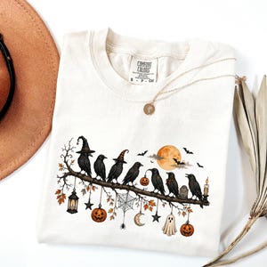 May include: A cream-colored t-shirt featuring a graphic of black crows perched on a branch. The design includes a full moon, bats, pumpkins, a spider web, a ghost, and a lantern. The shirt has a Comfort Colors label at the neck and a round neckline.