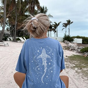 Puede incluir: Camiseta azul claro con un dise&ntilde;o de tibur&oacute;n ballena blanco y estrellas. El gr&aacute;fico est&aacute; en la parte posterior de la camiseta. La camiseta tiene un corte holgado. Escena de playa con palmeras al fondo.