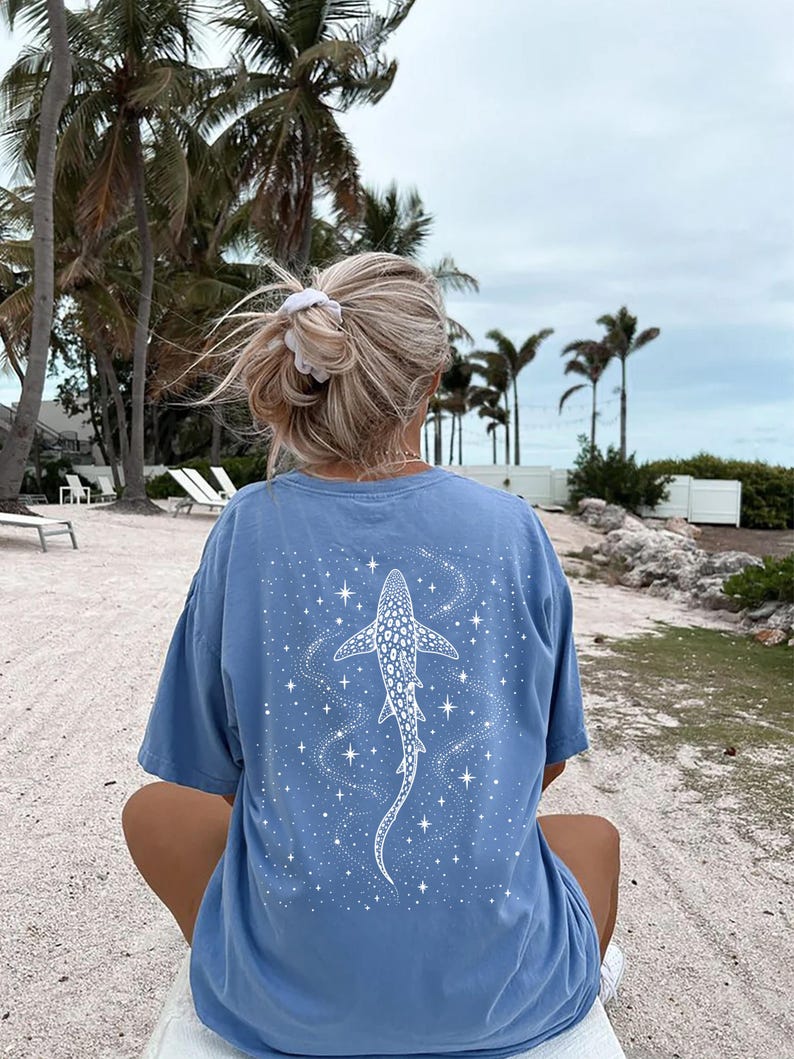 Puede incluir: Camiseta azul claro con un dise&ntilde;o de tibur&oacute;n ballena blanco y estrellas. El gr&aacute;fico est&aacute; en la parte posterior de la camiseta. La camiseta tiene un corte holgado. Escena de playa con palmeras al fondo.