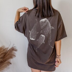 May include: Dark brown t-shirt featuring a white illustration of three whale sharks. The design includes detailed patterns of white spots on the whale sharks. The shirt has short sleeves and a relaxed fit.