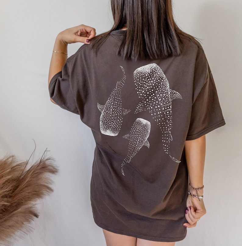 May include: Dark brown t-shirt featuring a white illustration of three whale sharks. The design includes detailed patterns of white spots on the whale sharks. The shirt has short sleeves and a relaxed fit.