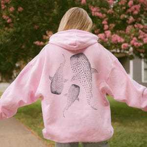 May include: A light pink hoodie featuring three black and white whale shark illustrations on the back. The whale sharks are detailed with dotted patterns. The hoodie has a hood and long sleeves.