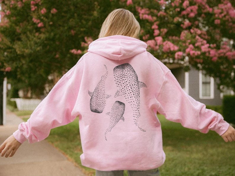May include: A light pink hoodie featuring three black and white whale shark illustrations on the back. The whale sharks are detailed with dotted patterns. The hoodie has a hood and long sleeves.
