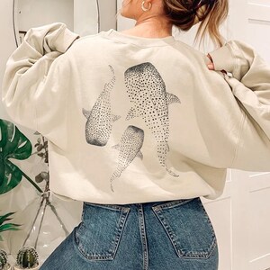 May include: Beige sweatshirt with a whale shark design on the back. The design features three whale sharks in a black dotted pattern. The sweatshirt has long sleeves and a crew neck. The model is wearing blue jeans.