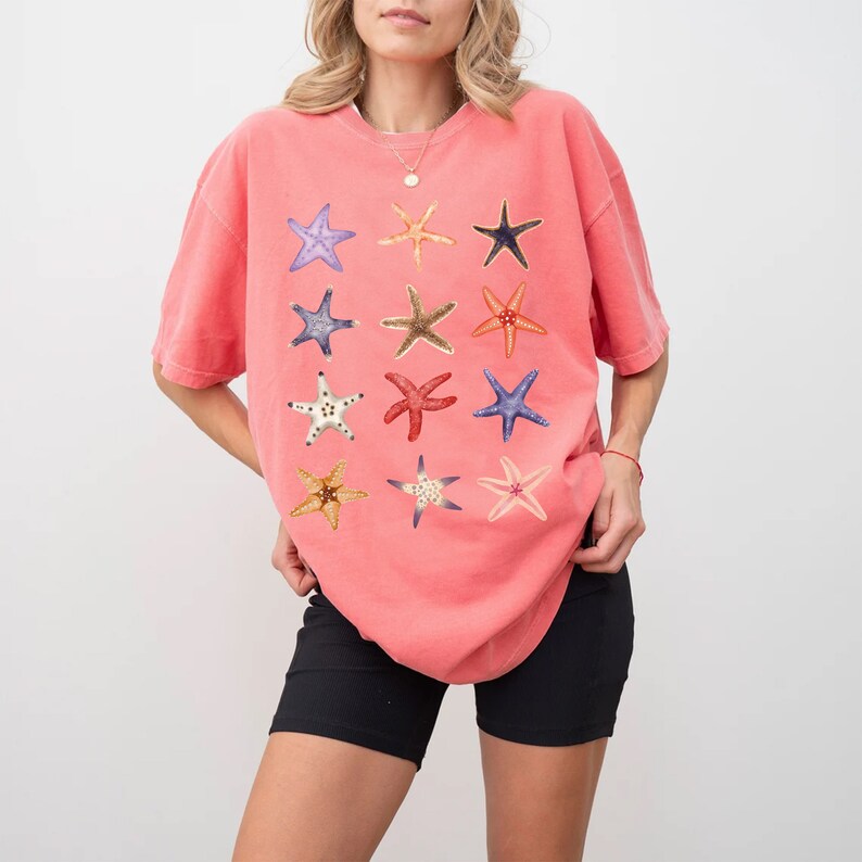 May include: A coral pink oversized t-shirt with a graphic print of 12 colorful starfish. The shirt has a crew neck and short sleeves.